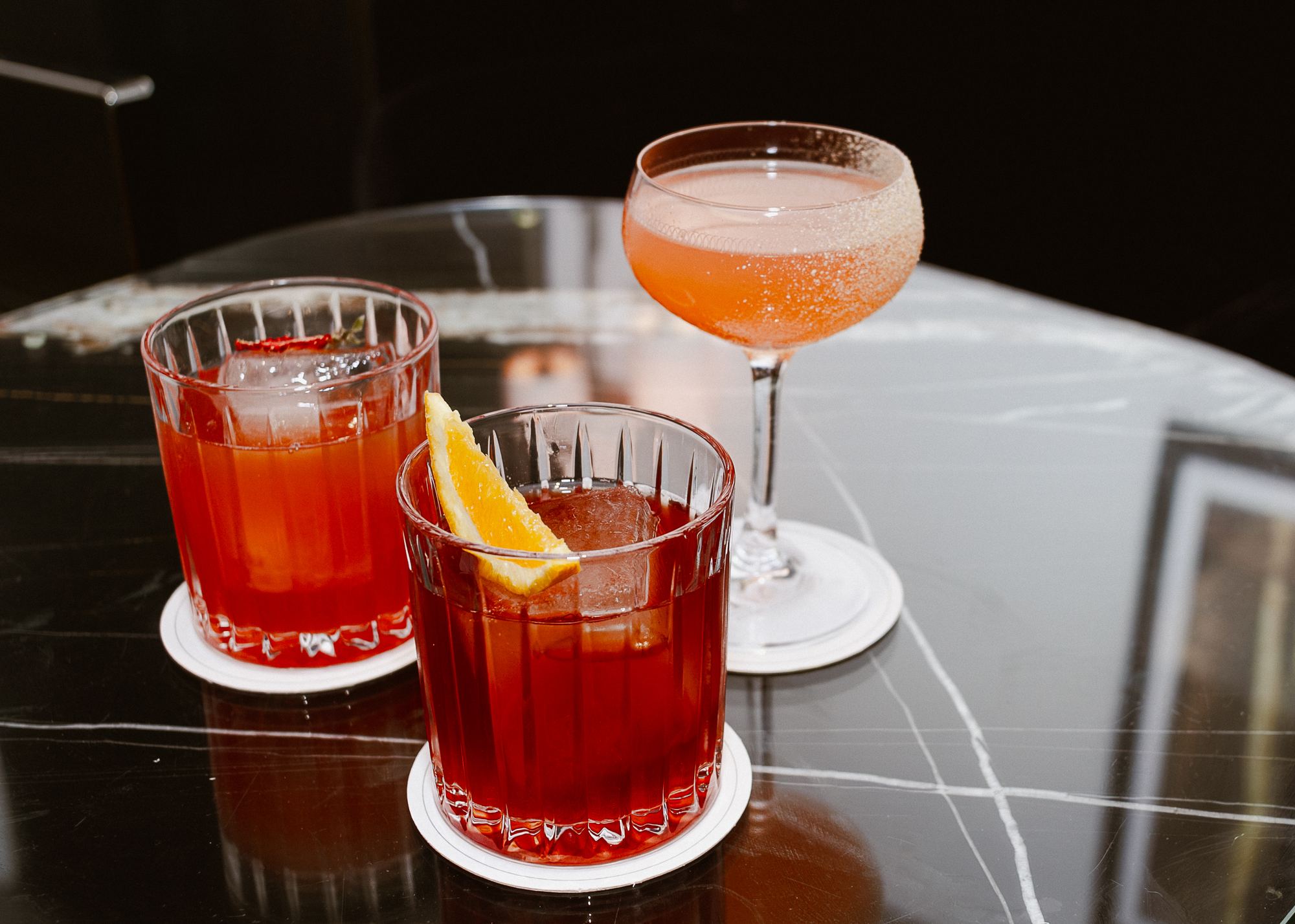 Two cocktails garnished with citrus on a dark bar top